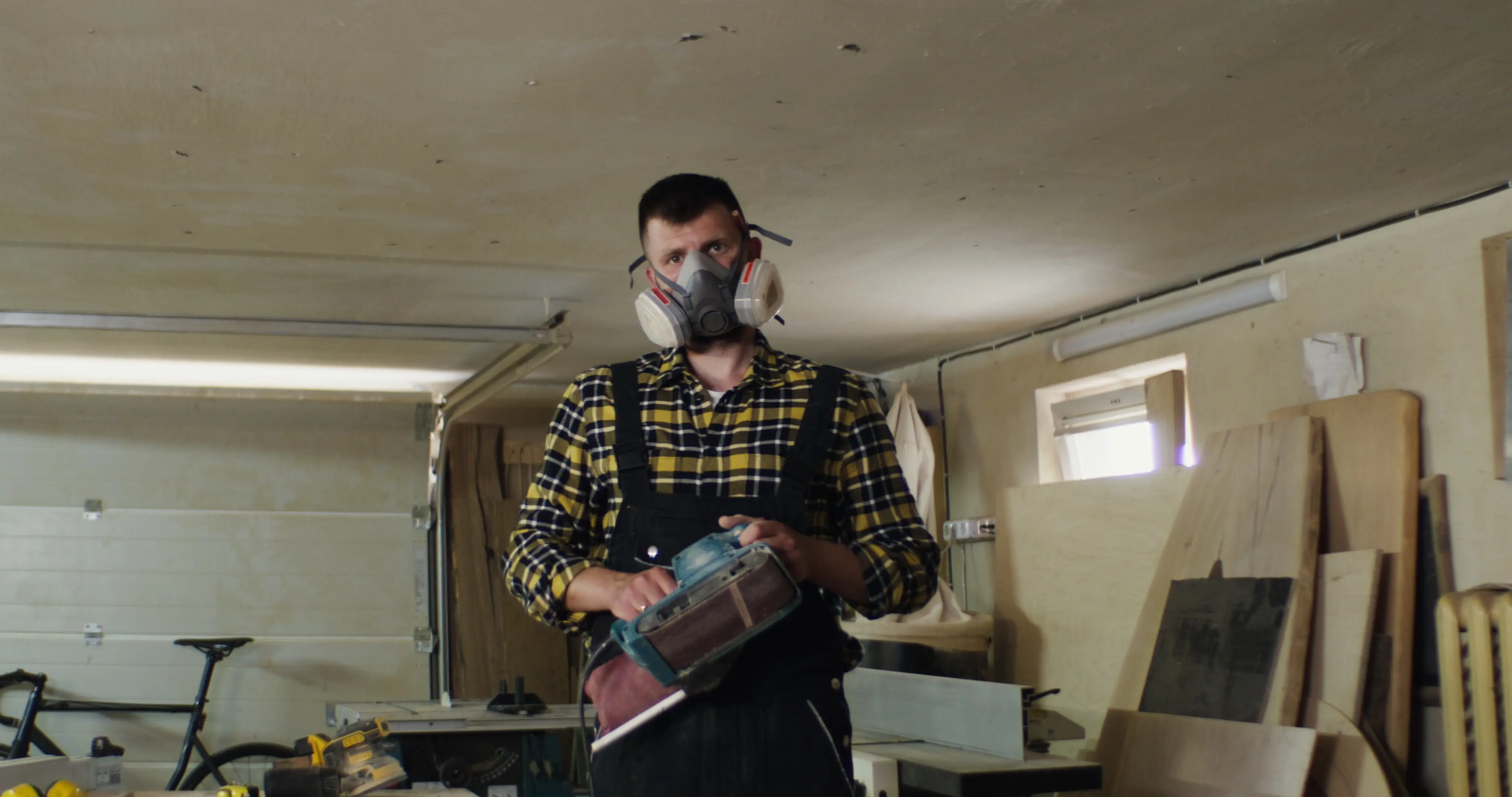 A man in a protective mask with a woodworking tool looks at the camera.